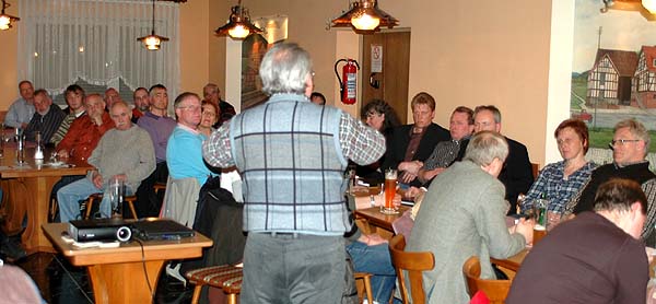 Männer und Frauen sitzen an Holztischen und hören aufmerksam zu 